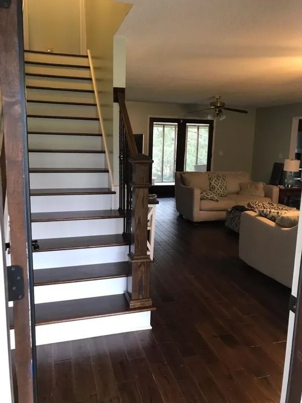 Living room with staircase, dark wood floors, beige sofas, and a windowed seating area.