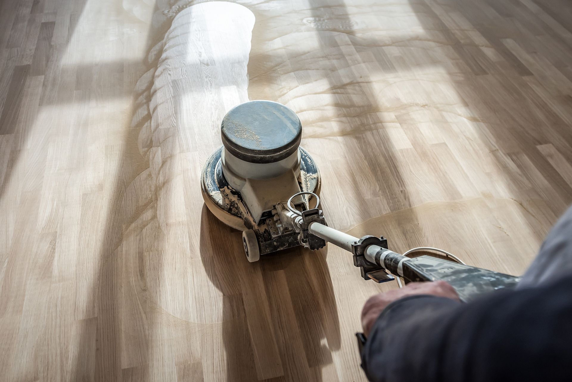 Person sanding a wooden floor with a floor sander in a sunlit room