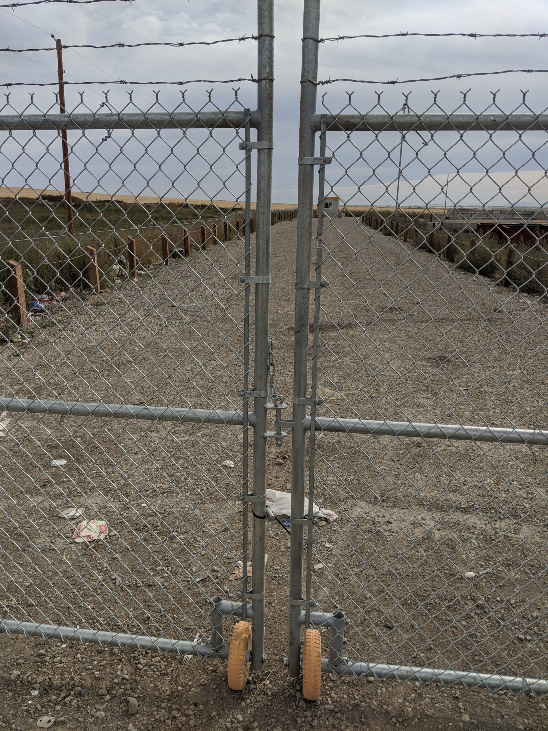 Two chain link gates, closed, with wheels on a gravel path, under barbed wire and a cloudy sky.
