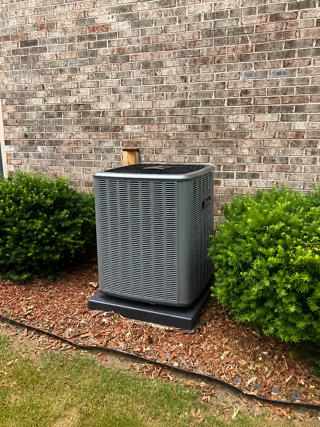 Outdoor air conditioner unit beside a brick house wall and shrubs in a landscaped yard