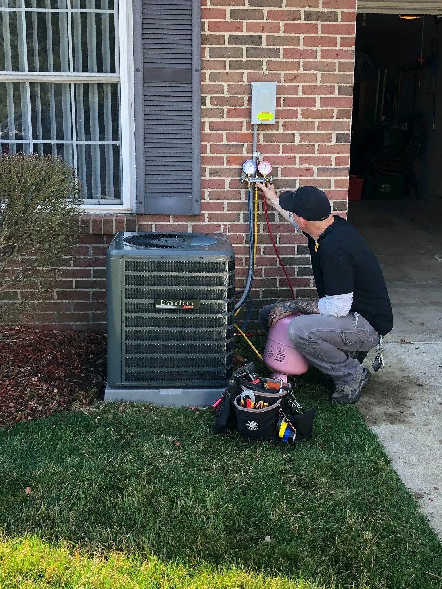 Technician kneeling beside a brick house, inspecting an outdoor HVAC unit with tools nearby.