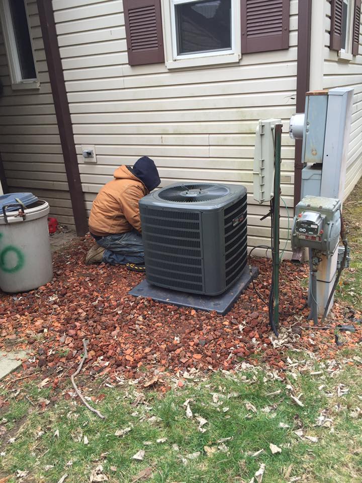Technician kneeling beside outdoor air conditioner unit next to house wall, inspecting equipment.