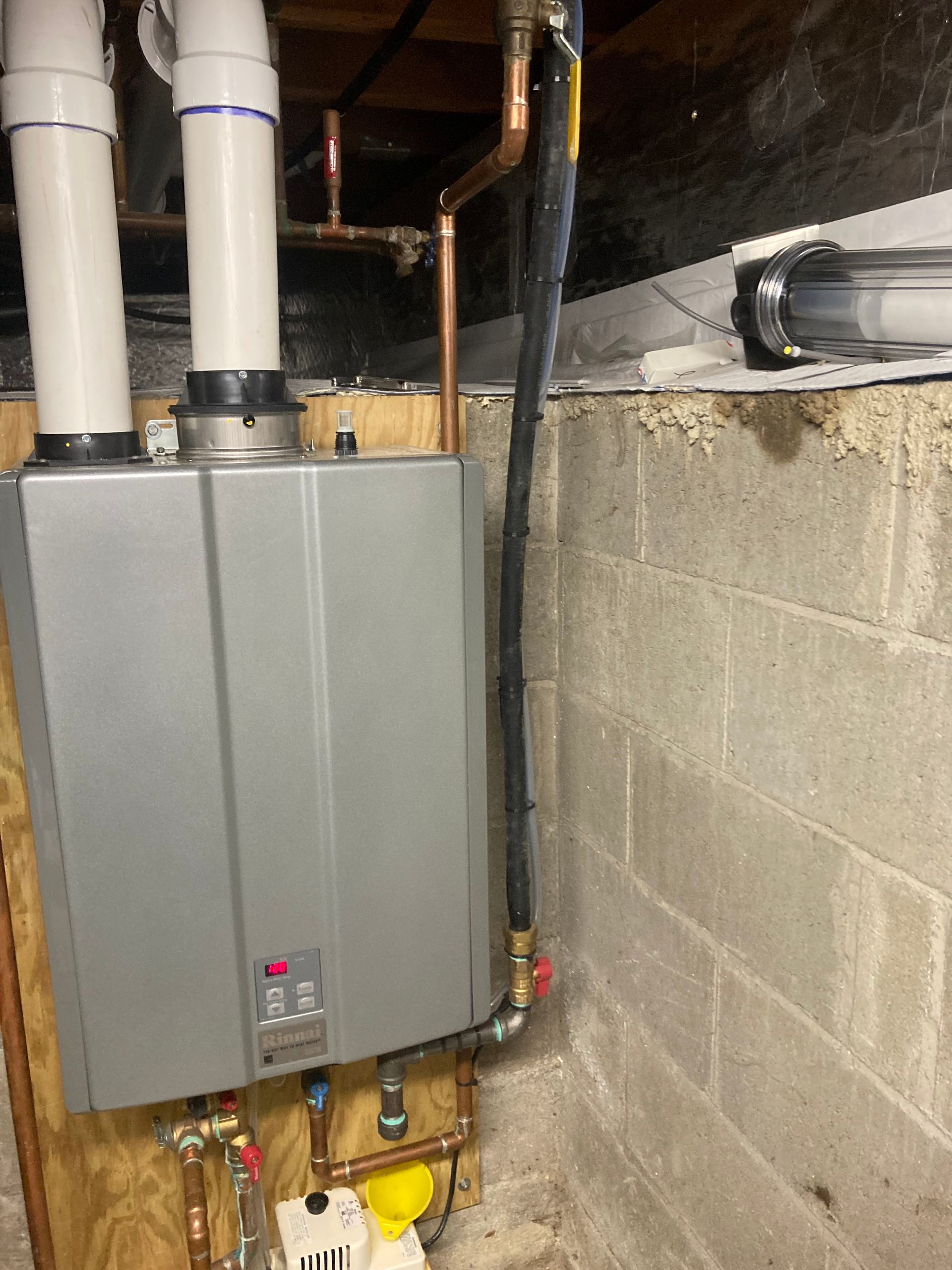 Wall-mounted gray water heater in a basement with white pipes and exposed cinder block walls.