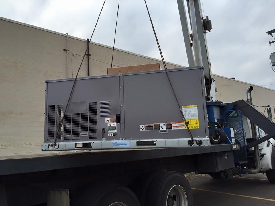 Gray industrial generator hoisted by crane onto a flatbed truck beside a building