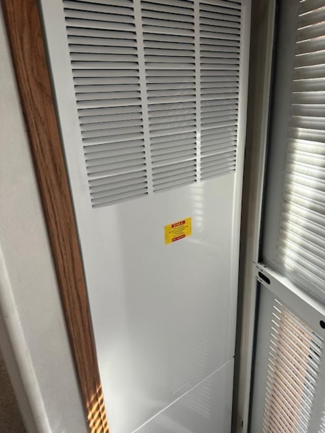 White louvered vent panel in a narrow closet or utility space, with a small yellow warning label.