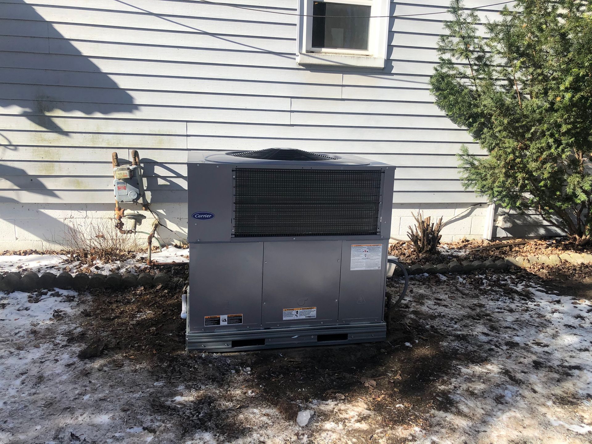 Outdoor HVAC unit beside a white house with snow on the ground and a tree to the right