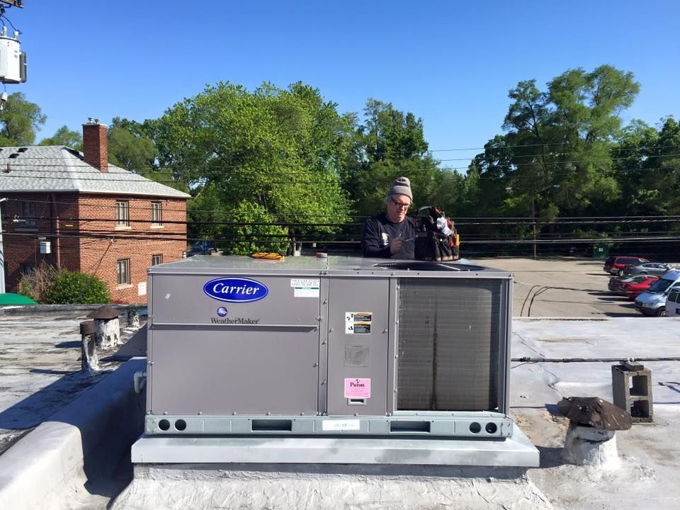 Gray Carrier rooftop HVAC unit on a flat roof, with a worker standing behind it under a blue sky.