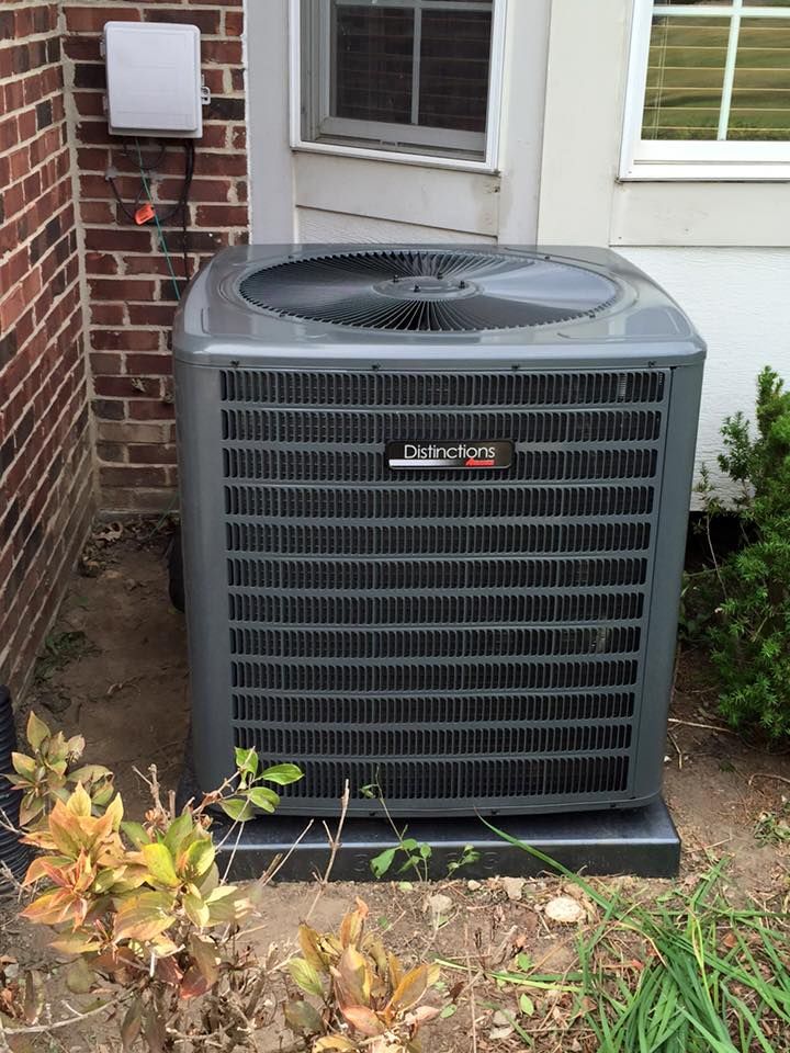 Outdoor air conditioner unit beside a brick house wall and window, with plants in front.