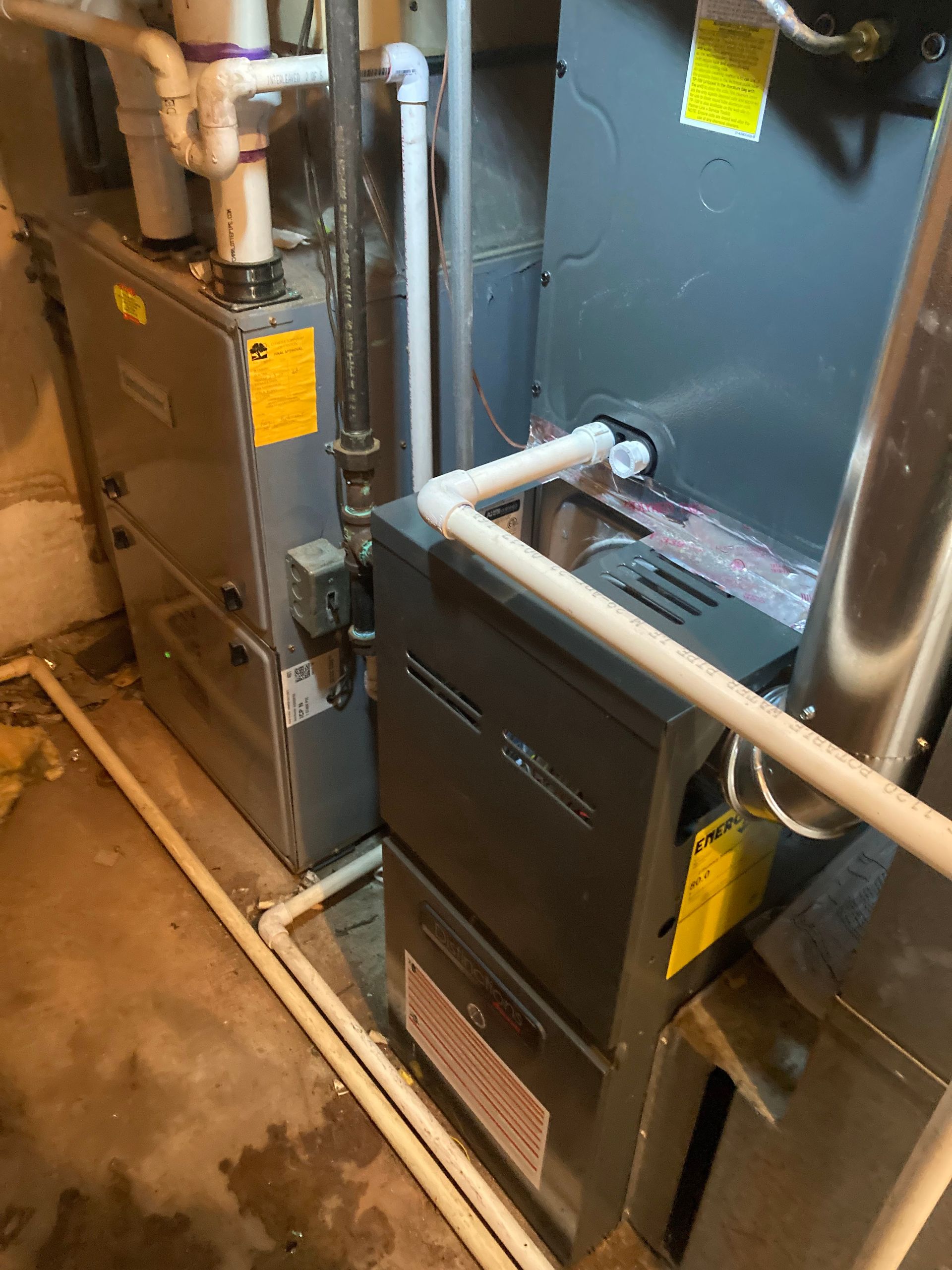 Two gray HVAC furnace units in a utility closet with white pipes and yellow labels.