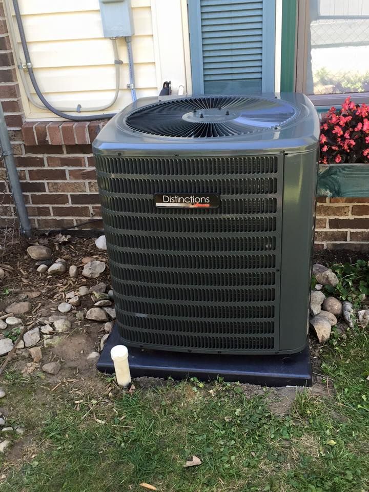 Outdoor central air conditioner unit beside a house wall and window, on grass and rocks.