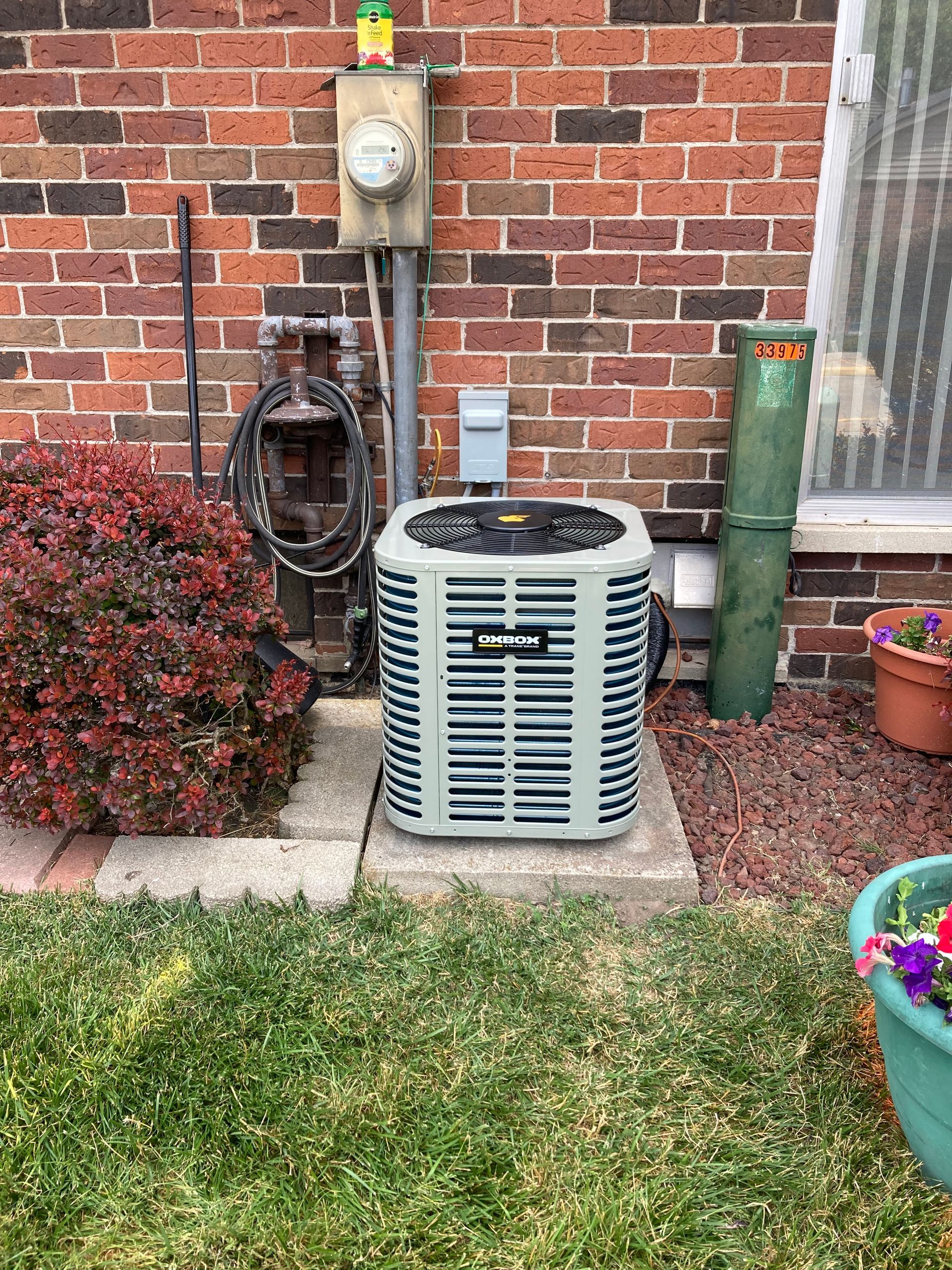 Outdoor HVAC unit beside a brick house wall with utility meter, hose, and flower beds.