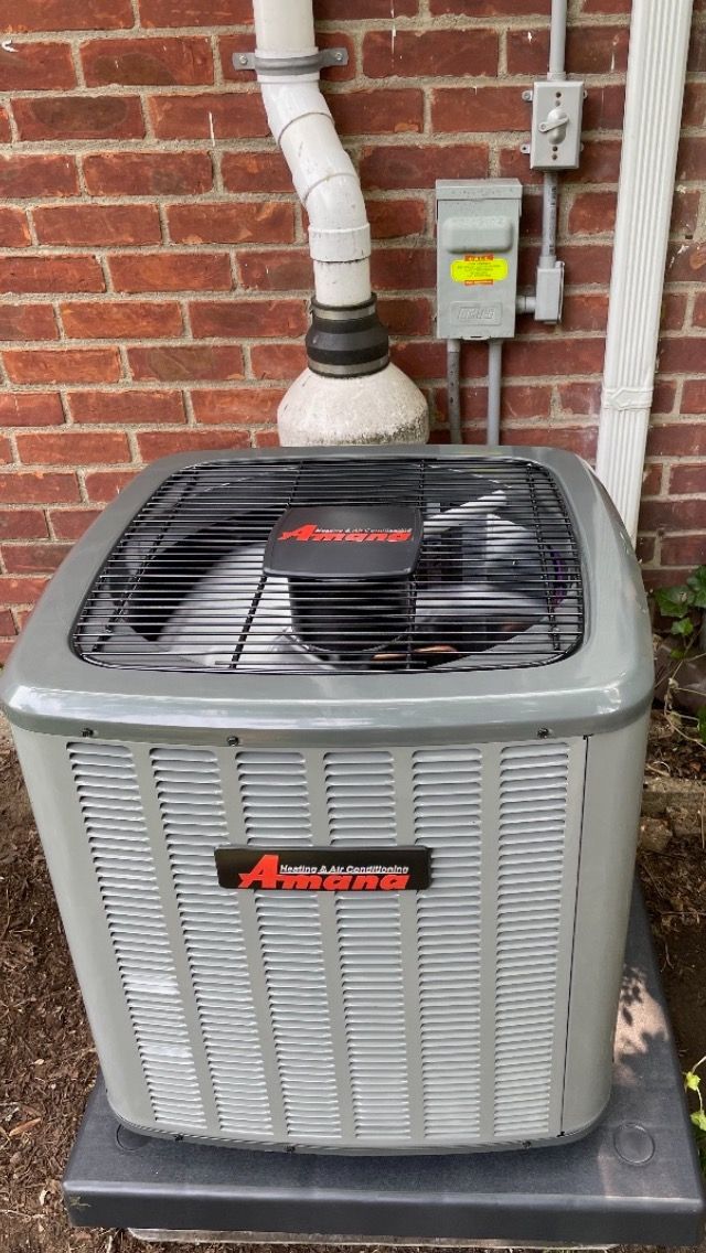 Gray Amana outdoor air conditioner unit beside a brick wall with white vent pipe and electrical box