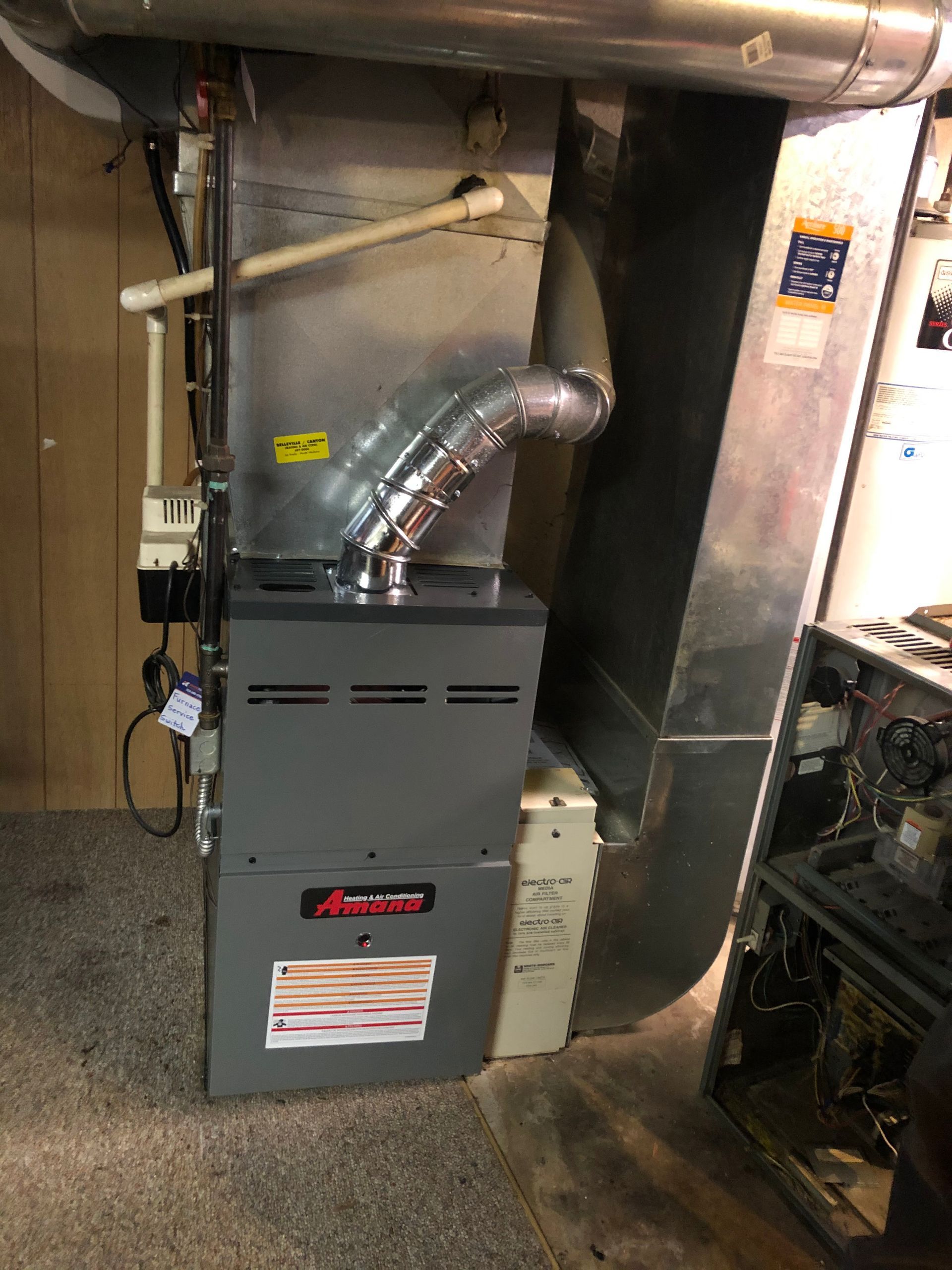 Basement furnace with silver ductwork and vent pipe next to a large metal HVAC unit