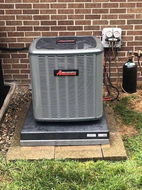 Outdoor central air conditioner unit beside a brick wall on a concrete pad with electrical equipment nearby