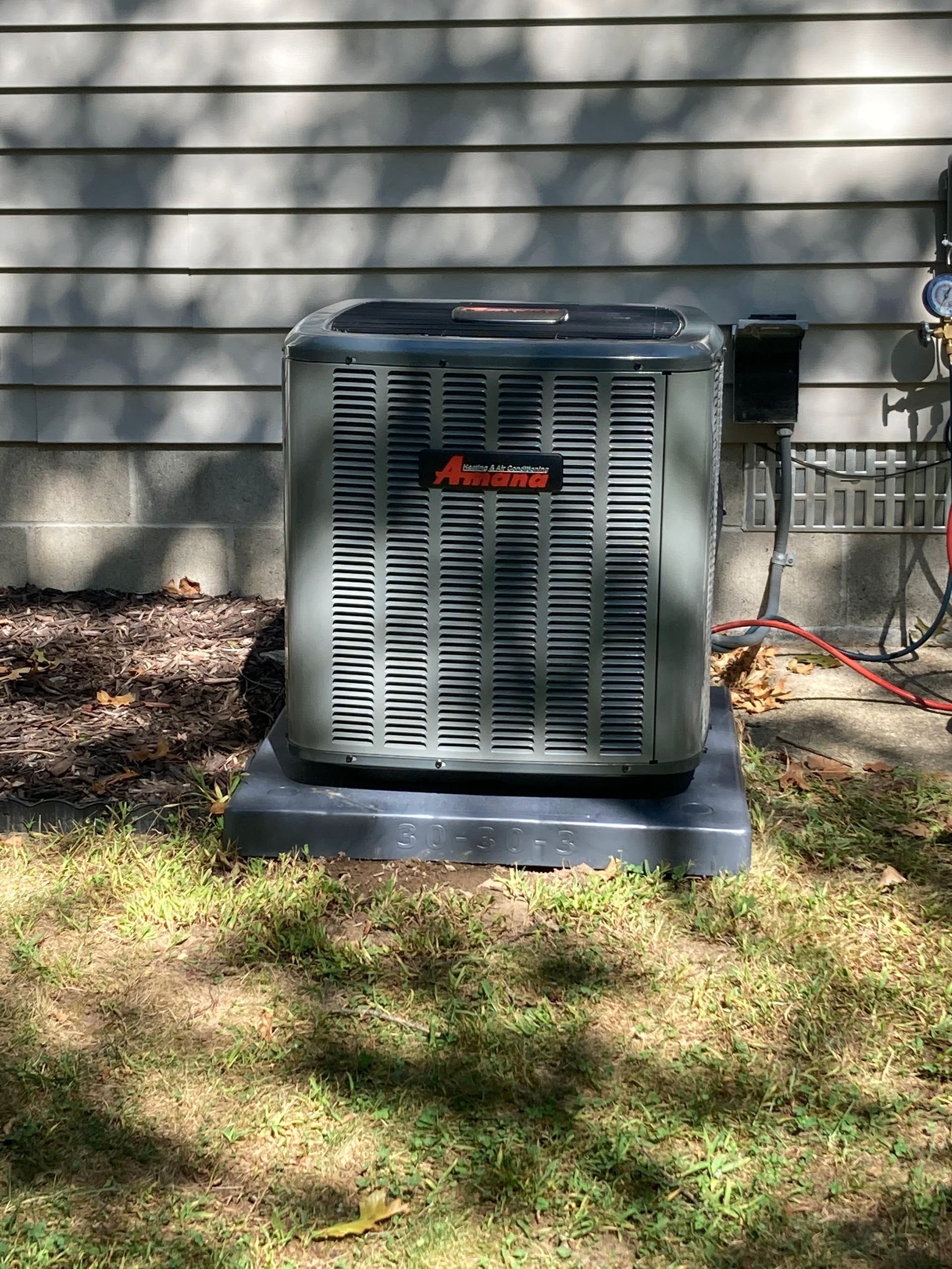 Outdoor HVAC unit on a concrete pad beside a house wall, with grass and leaves in the yard.