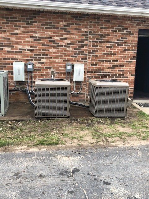 Two outdoor HVAC units beside a brick wall with utility meters and cables.