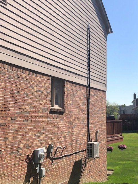Side of a brick house with tan siding, utility meters, a window, and a grassy yard under blue sky.