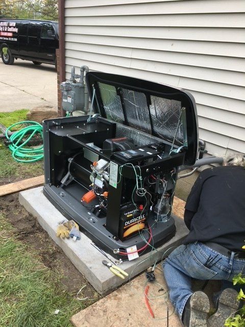 Open control box and machinery on a concrete pad beside a house, with a person kneeling nearby and tools on the ground