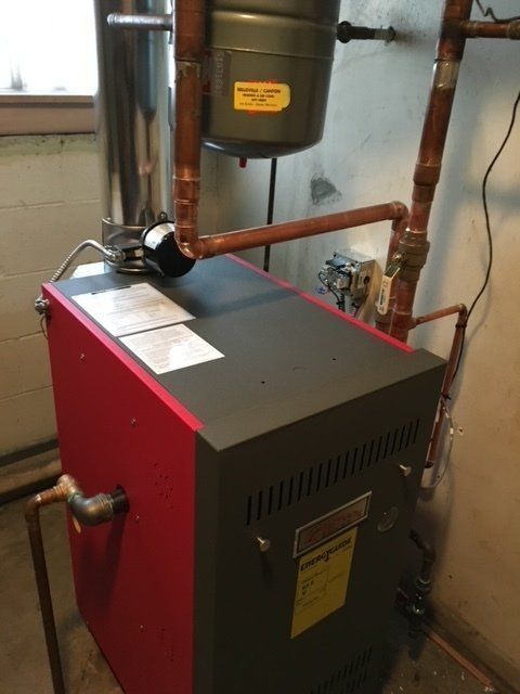 Red boiler unit with copper pipes and a small expansion tank in a basement utility room