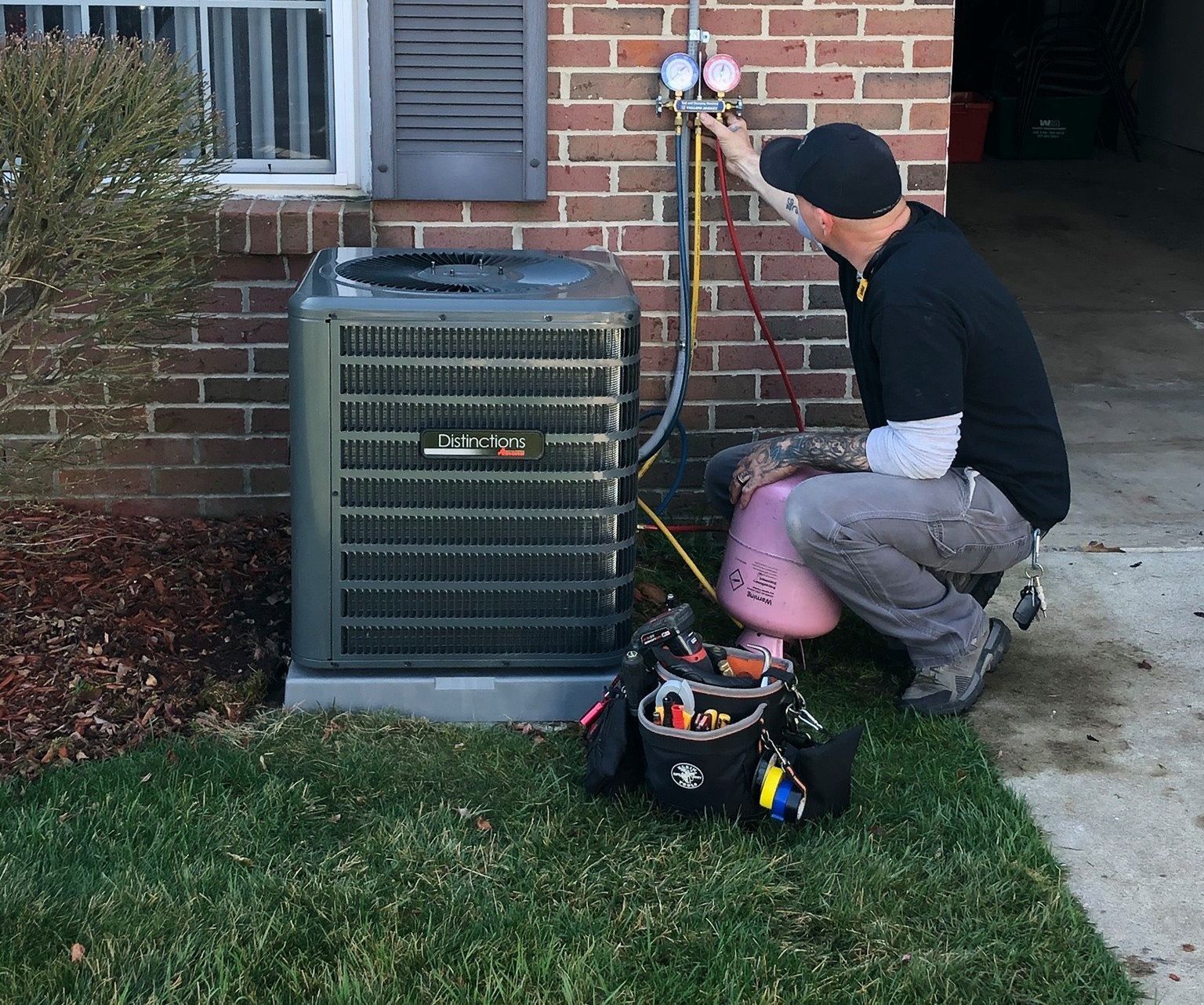 Technician kneels beside an outdoor HVAC unit, inspecting it with tools next to a brick house.
