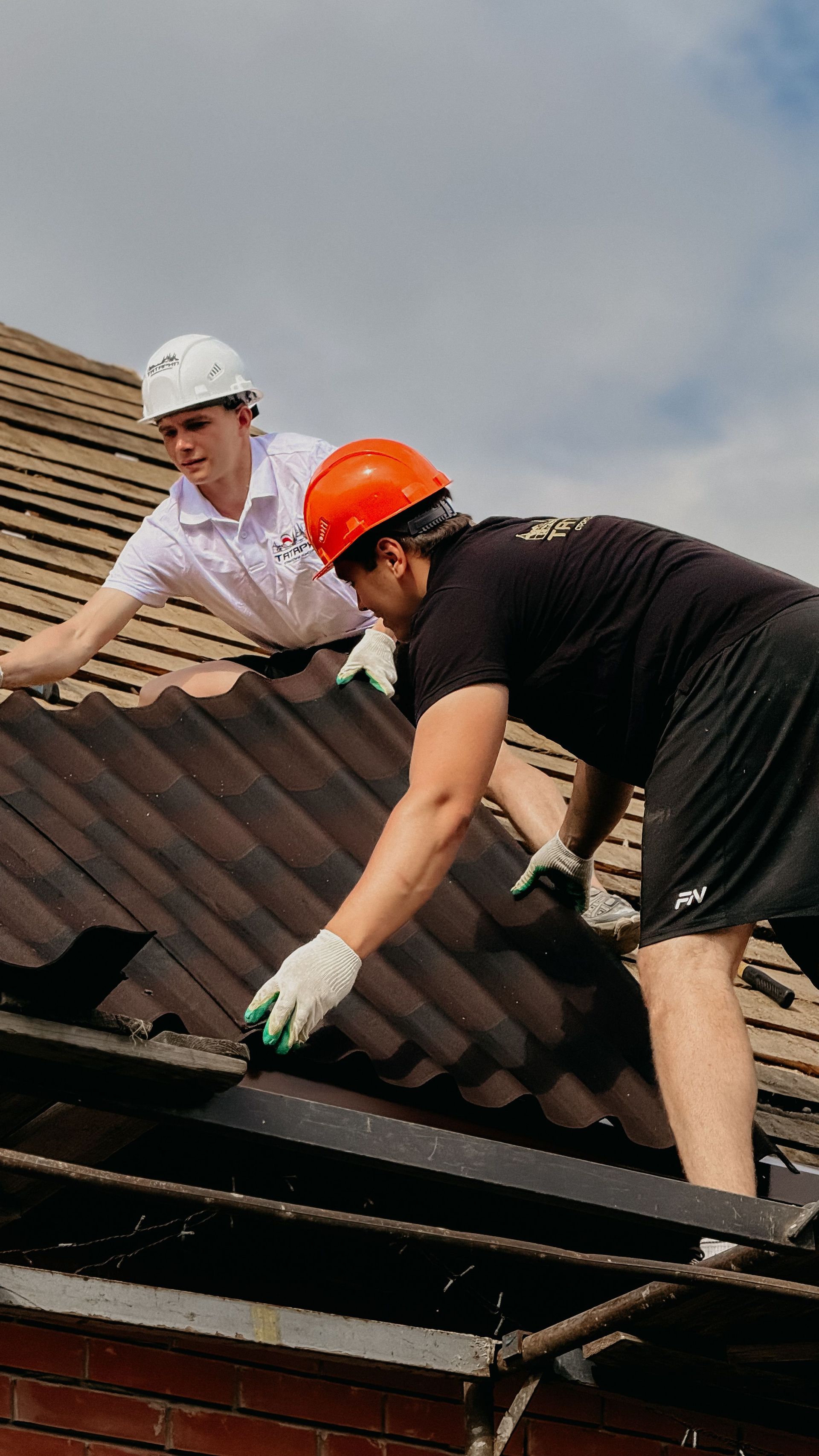 Two roofers in helmets repairing brown roof shingles on a house under a cloudy sky