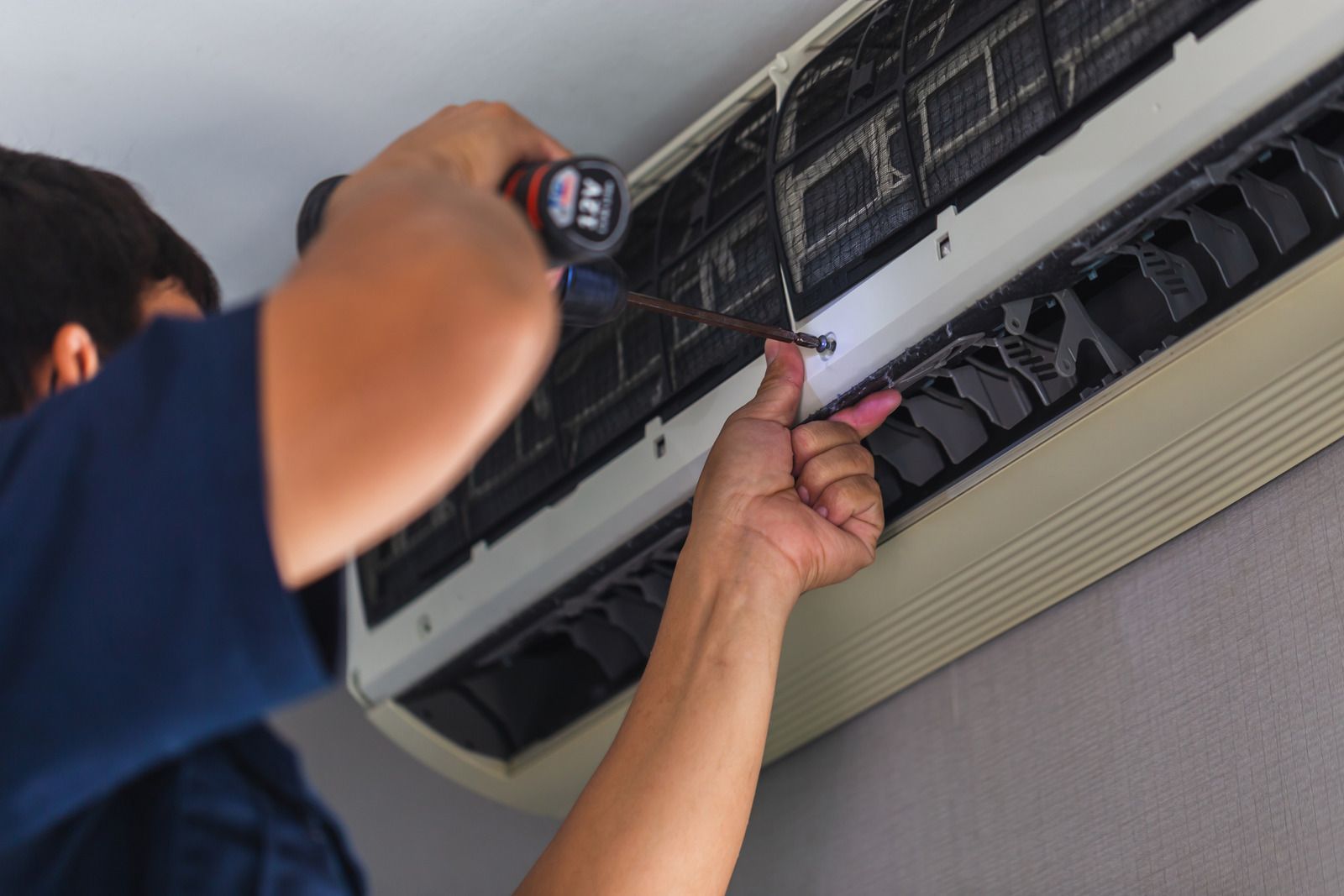 Technician cleaning a wall-mounted air conditioner filter with a screwdriver and flashlight