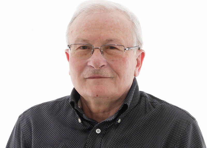 An older man wearing glasses and a black shirt is standing in front of a white background.
