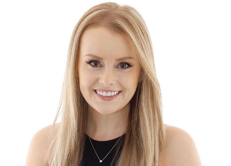 A woman with long blonde hair is smiling and wearing a black top and necklace.
