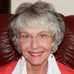 A woman wearing glasses and a red jacket is smiling while sitting in a chair.