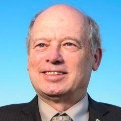 A bald man in a suit and tie is smiling in front of a blue sky.