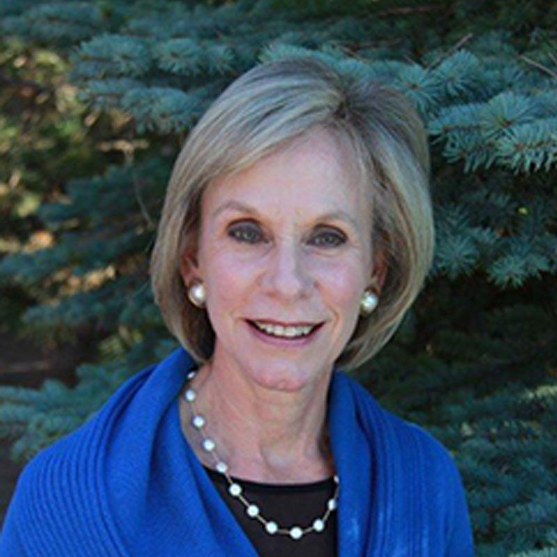 A woman wearing a blue sweater and pearls is smiling in front of a pine tree.