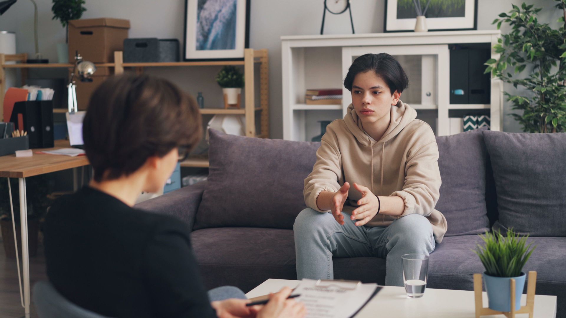 Two people in a living room during a discussion, one seated on a couch and one taking notes at a table.
