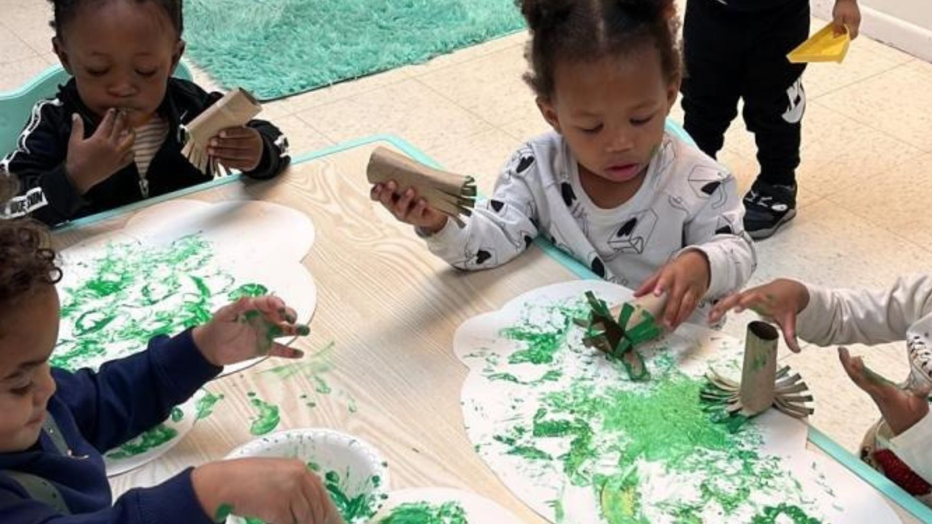 Children painting with green paint at a table, making handprints on white paper in a classroom.