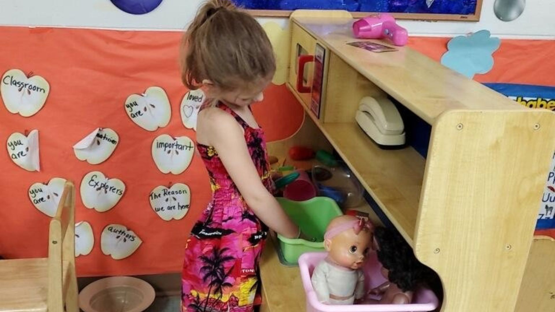 Child playing with dolls at a classroom play area, using a green scoop and pink bin.