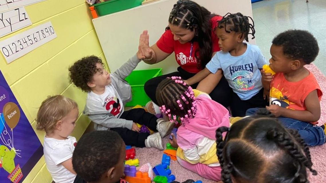 Children sit in a circle on a classroom rug, clapping and raising hands during group time.