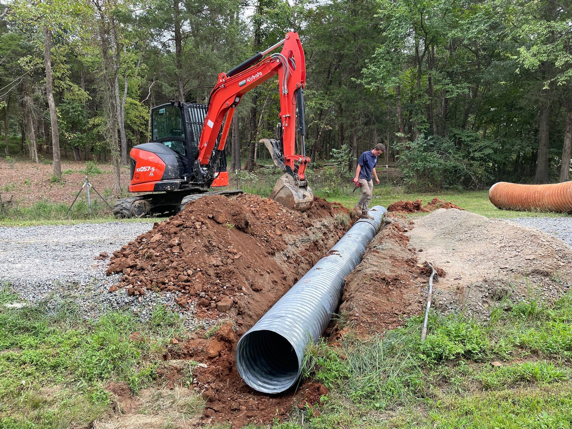 An excavator and a person are installing a corrugated metal drainage pipe in a trench outdoors.