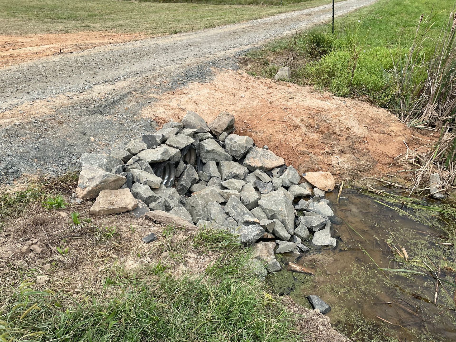 Pile of gray rocks in a ditch beside a gravel road and brown dirt patch. Green grass surrounds it.
