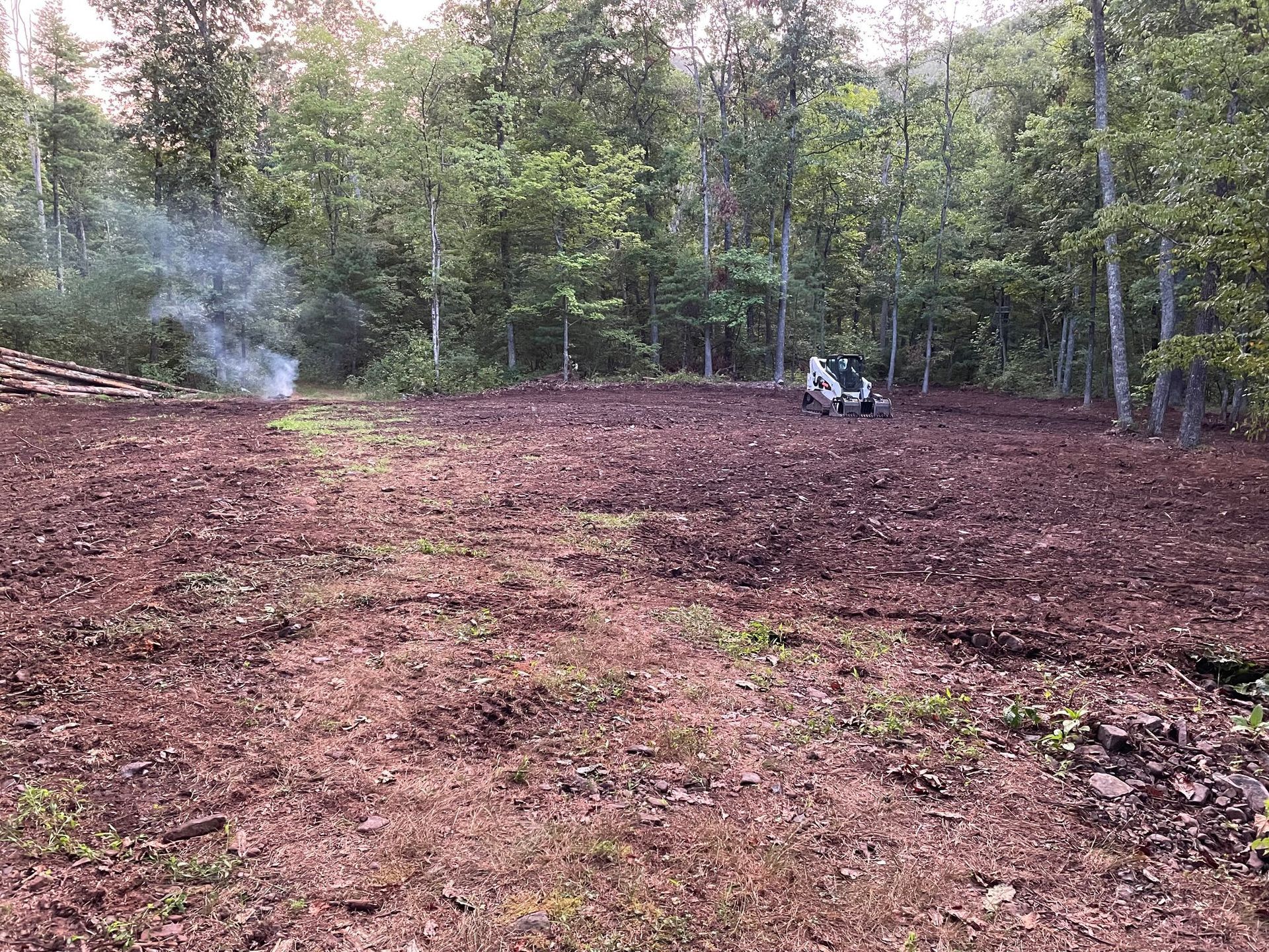 A cleared forest area with an excavator. Smoke rises near a pile of logs, brown soil.