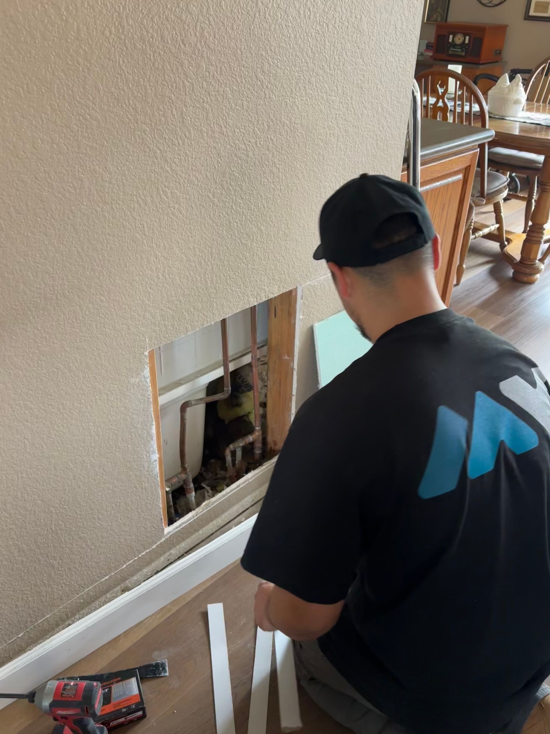 Worker repairing a wall cutout indoors, with tools and white trim pieces on the floor.