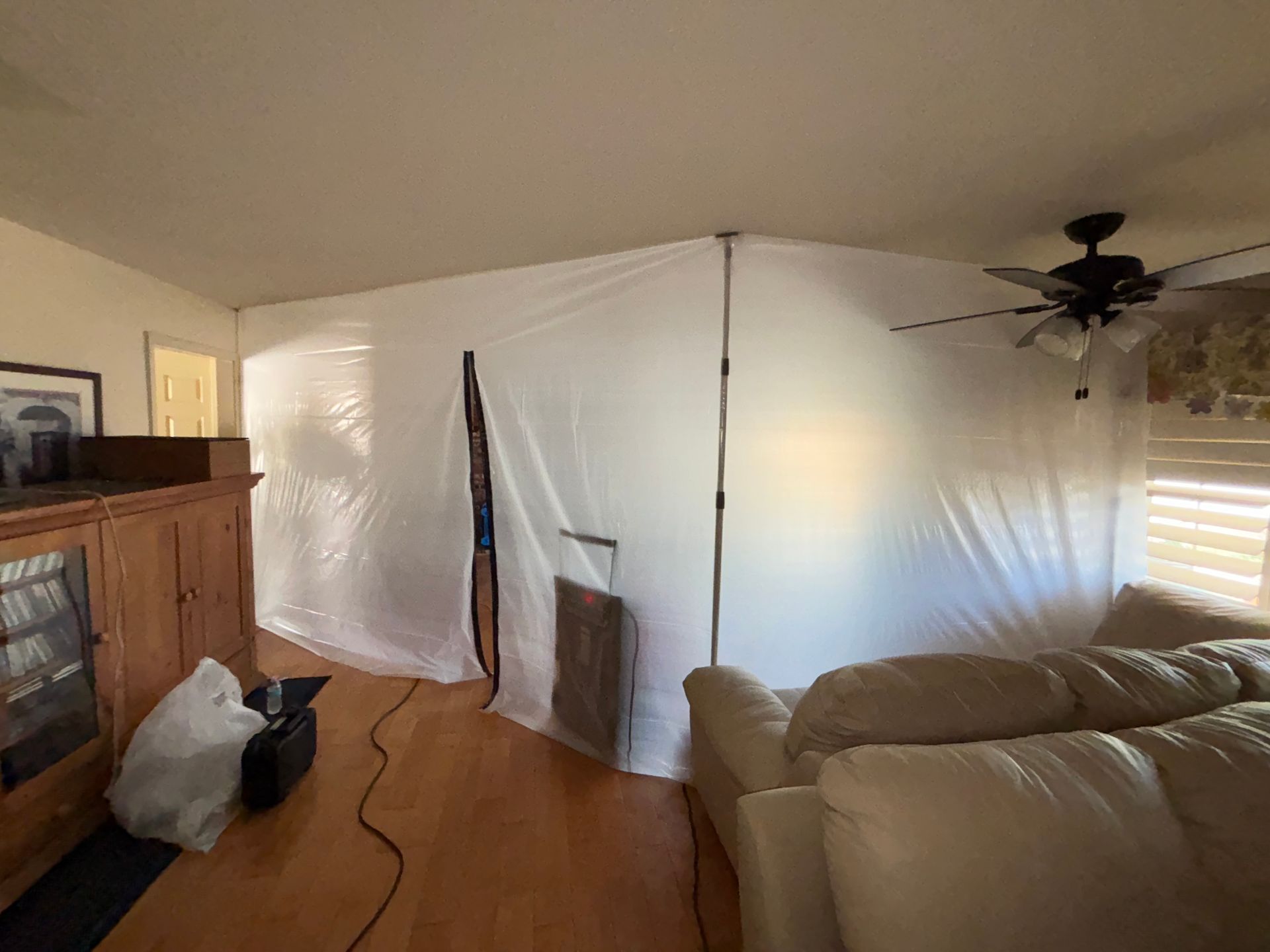 Living room corner with white plastic sheeting, wood floor, tan couch, TV cabinet, and ceiling fan