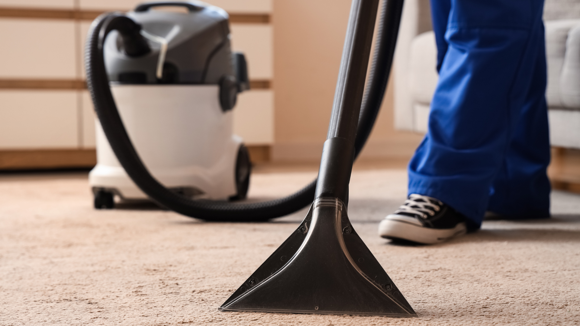 Vacuum cleaner on carpet beside a person in blue pants and sneakers, cleaning a room