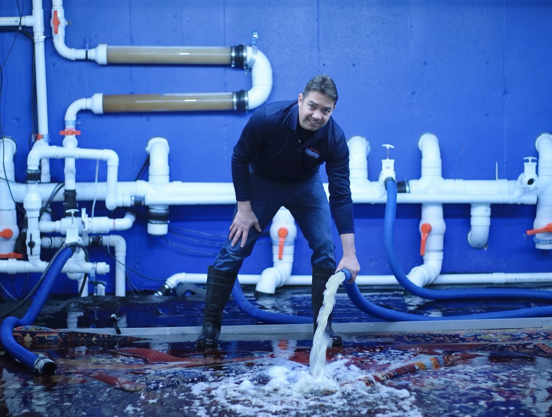 Man in dark clothes crouches by white pipes in a blue industrial room, handling water on the floor.