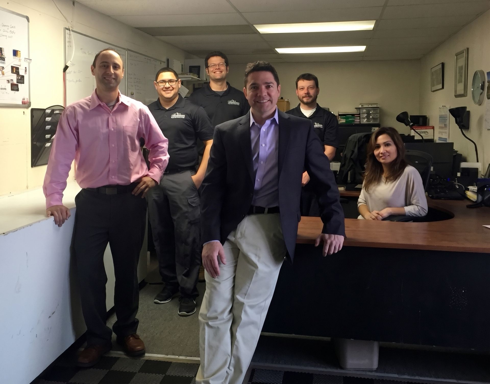 Five coworkers pose in an office hallway, with one man in a suit leaning on a desk in front.