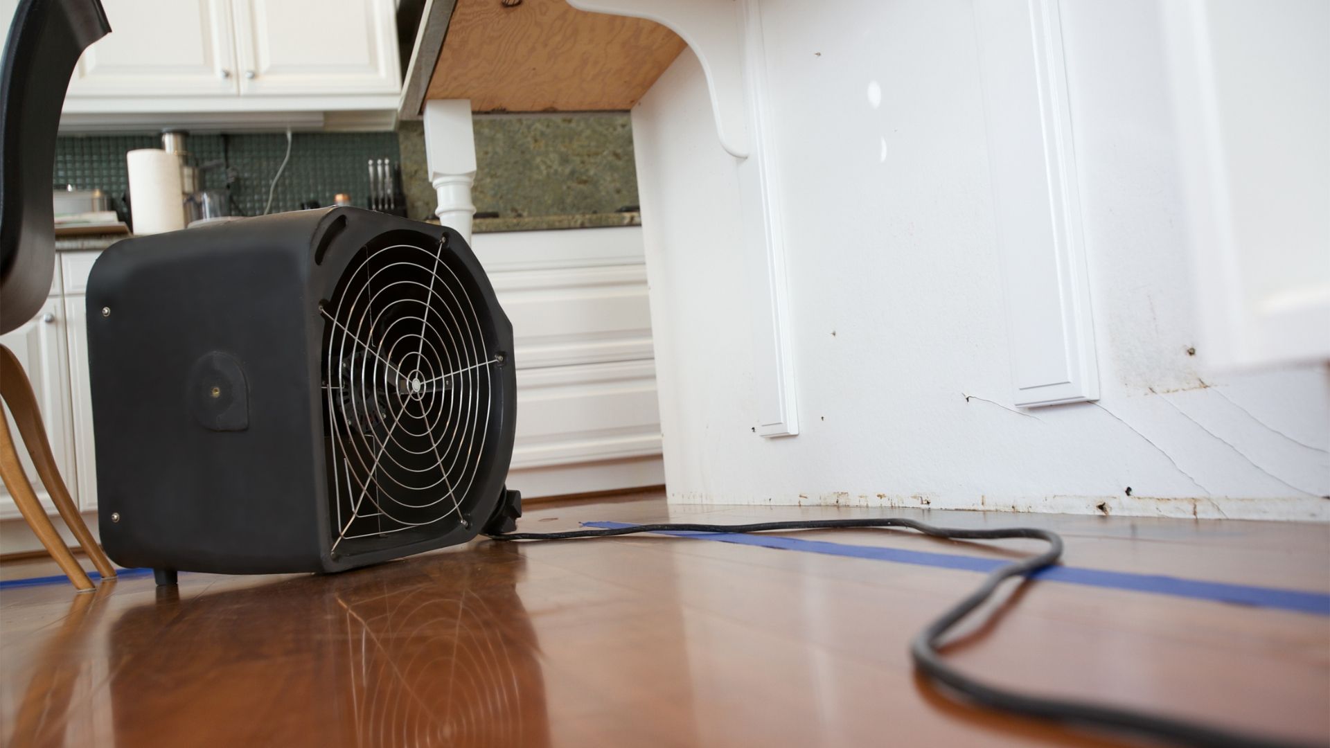 Black portable fan on a hardwood floor under a desk, with cables in a kitchen or office space.