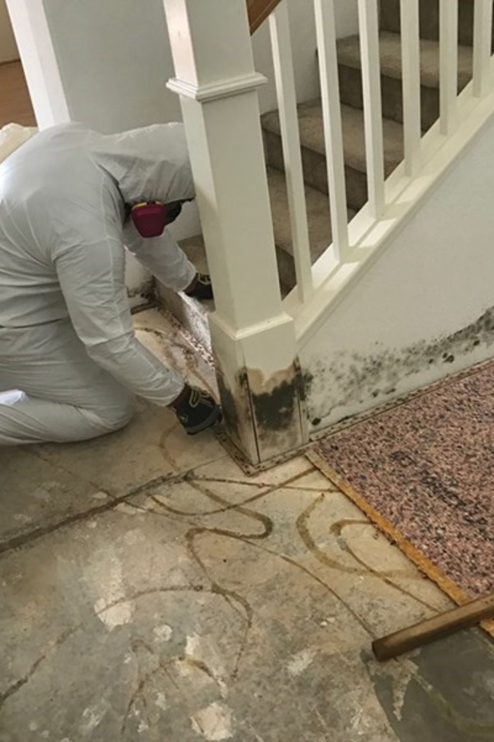 Worker in white coveralls inspecting mold or water damage at the base of a staircase.
