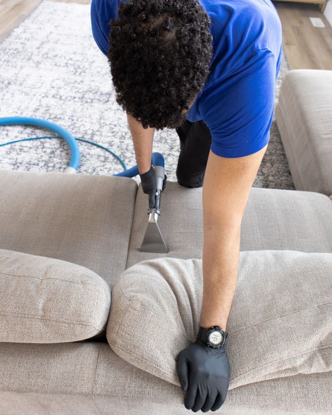 Person vacuuming a light gray couch with a handheld cleaner while wearing blue shirt and black gloves