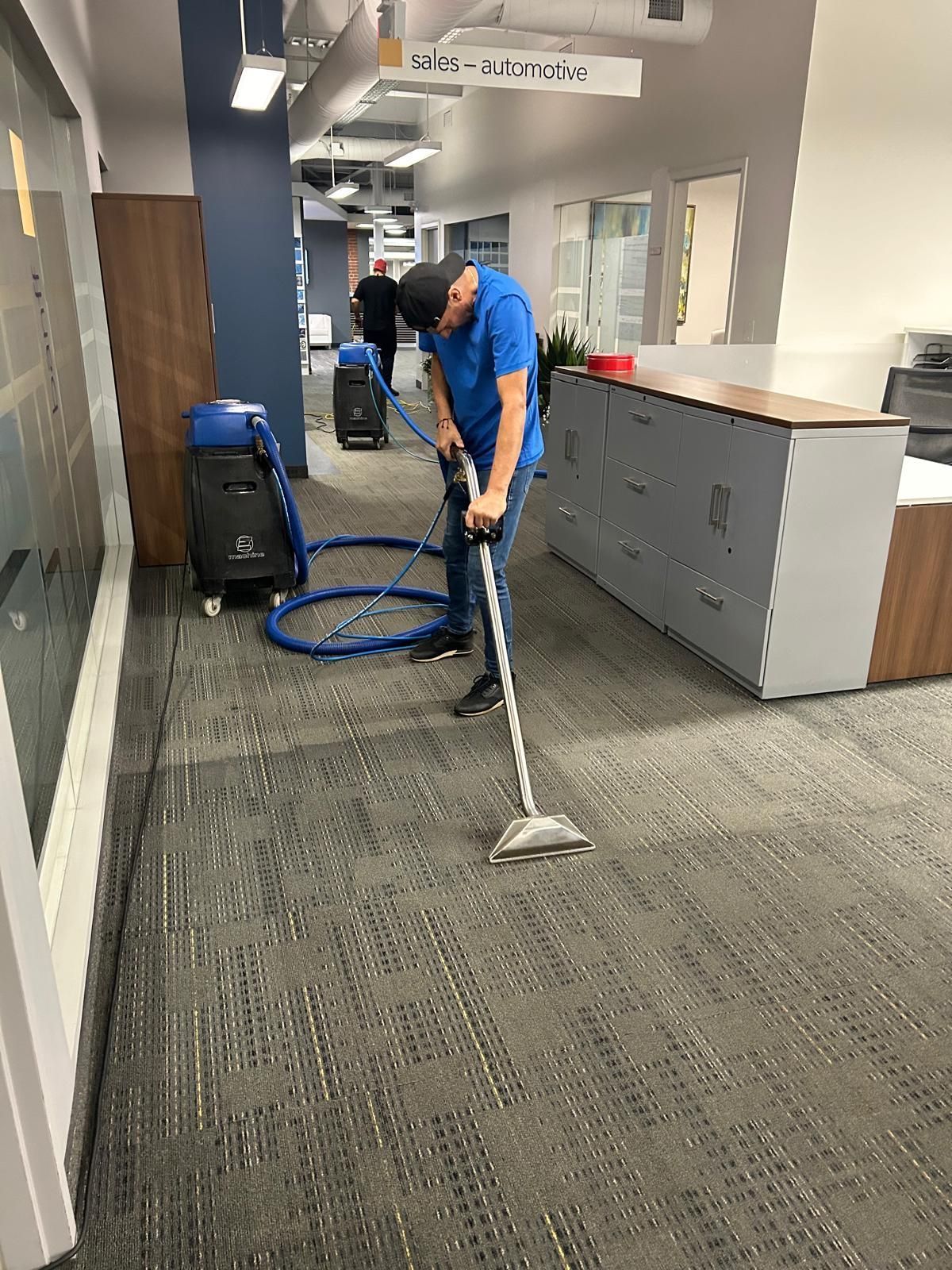 Worker cleaning carpet in an office hallway with a vacuum and blue hose