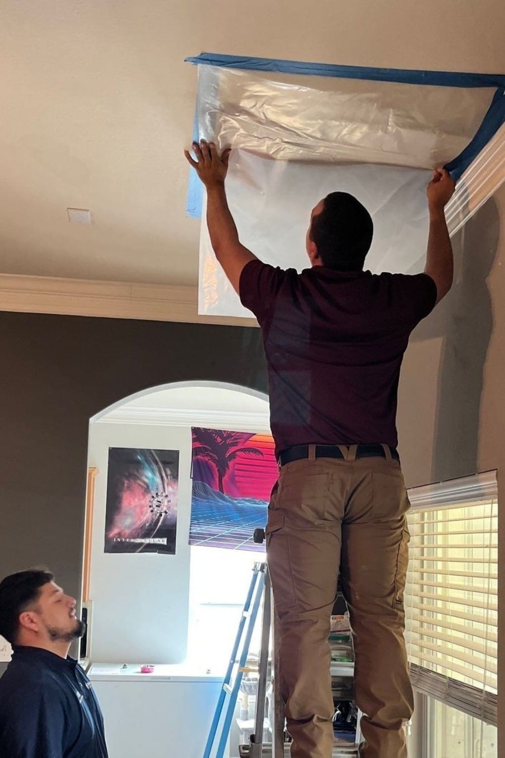 Man standing on a ladder taping plastic sheeting over a ceiling opening while another person watches