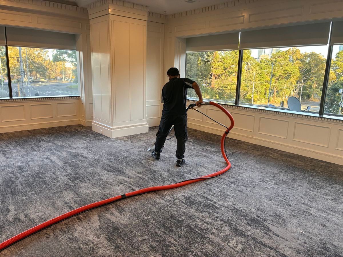 Worker vacuuming gray carpet in a large empty room with windows and white walls