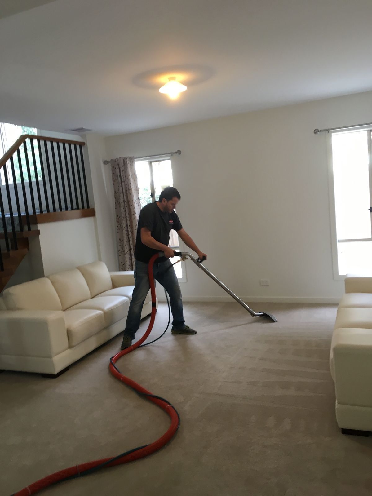 Person vacuuming beige carpet in a living room with white sofas and stairs.